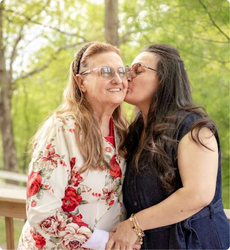 Senhora sorridente recebendo um beijo no rosto de uma mulher mais jovem, em um momento de carinho ao ar livre na fazenda